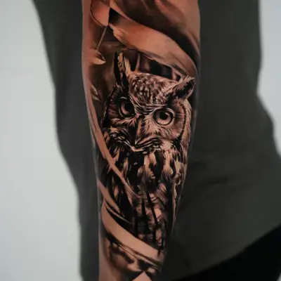 Black and grey realism tattoo of a great horned owl among tall reeds, wrapping a forearm.