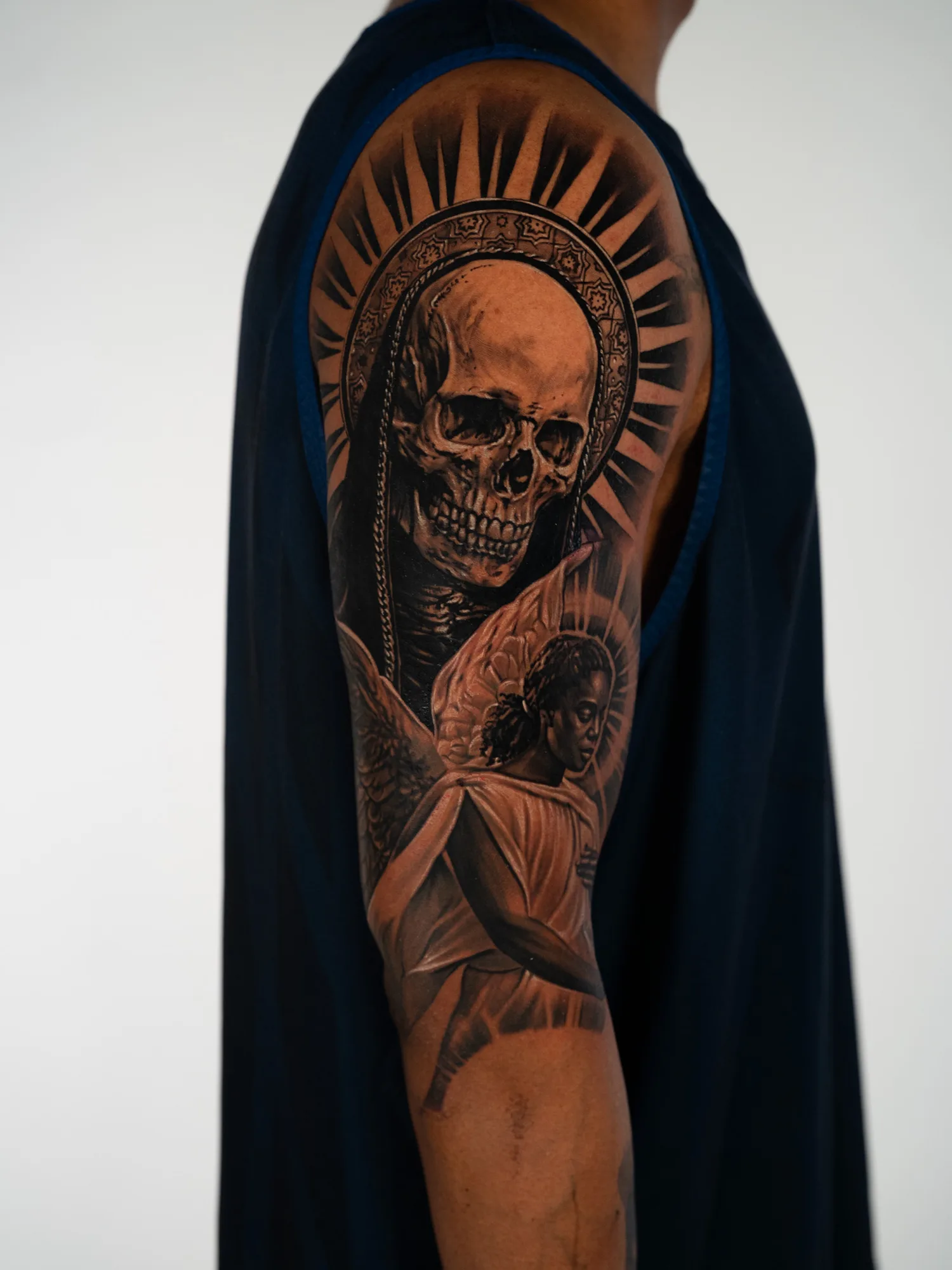 Black and grey realism tattoo in progress on a shoulder and upper arm, showing a skull in a hooded mantle under a rayed halo above a winged angel.