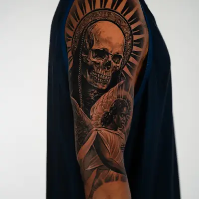 Black and grey realism tattoo in progress on a shoulder and upper arm, showing a skull in a hooded mantle under a rayed halo above a winged angel.