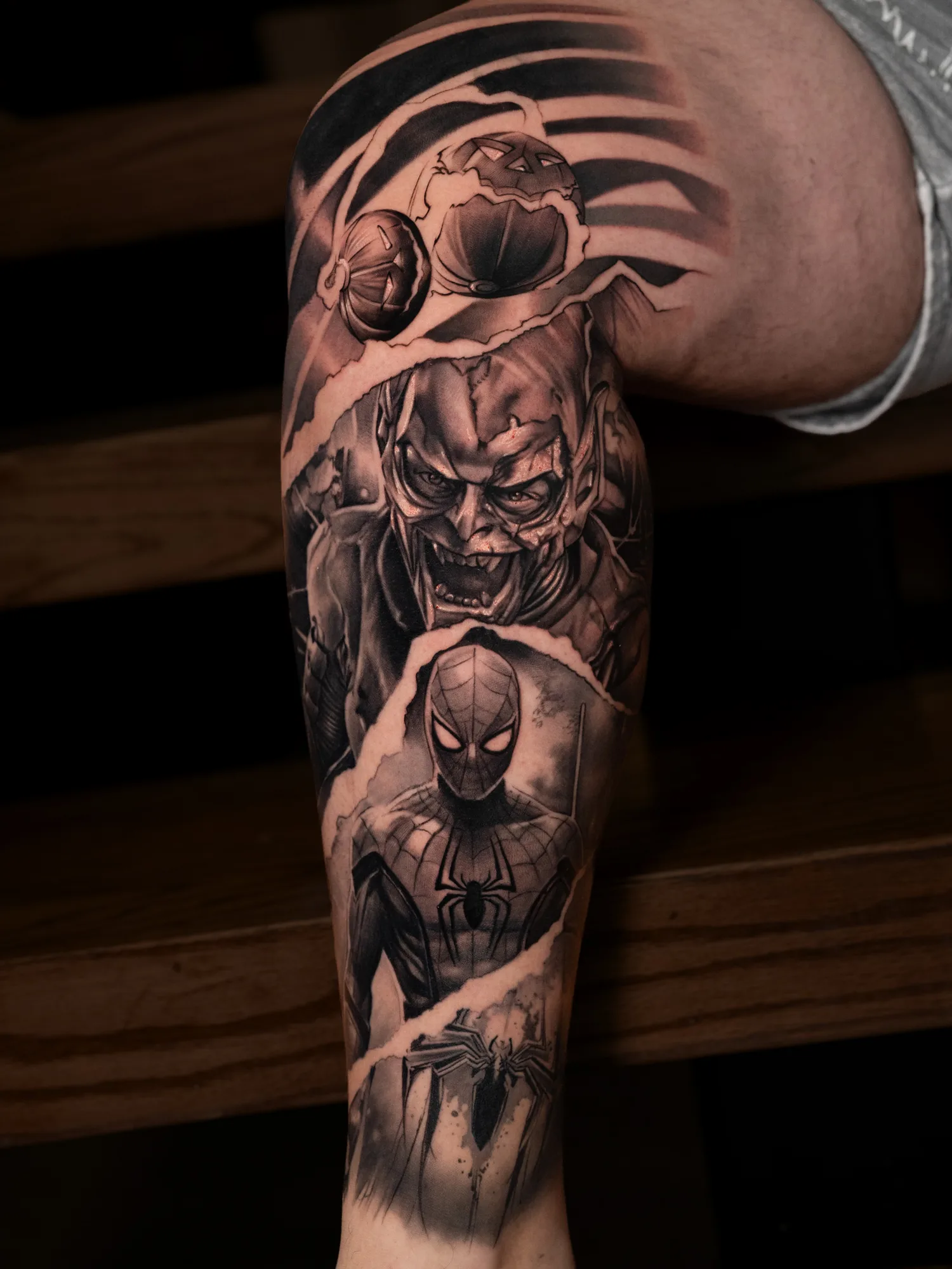 Black and grey realism tattoo down a lower leg with pumpkin bombs at the top, the Green Goblin in the middle and Spider-Man below, divided by torn paper edges.