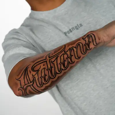 Black and grey tattoo of the name Autumn in ornate script with fine filigree, running from elbow to wrist along an outer forearm.