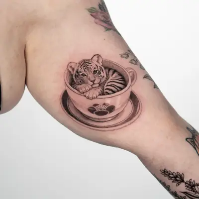 Black and grey tattoo of a tiger cub sitting in a teacup on a saucer, with a paw print on the side of the cup, on an inner bicep.