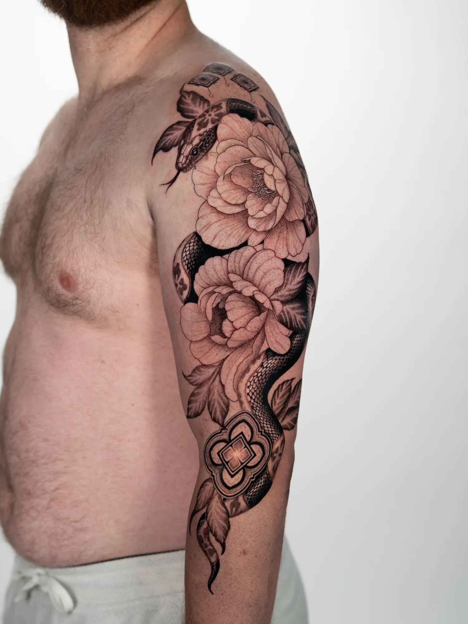 Black and grey half sleeve of a snake winding through two large peonies across a shoulder and upper arm, with ornamental medallions near the elbow.