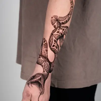 Black and grey tattoo of a viper with its head on the back of the hand and mouth open, its body patterned with geometric ornament, winding up the forearm to the elbow.