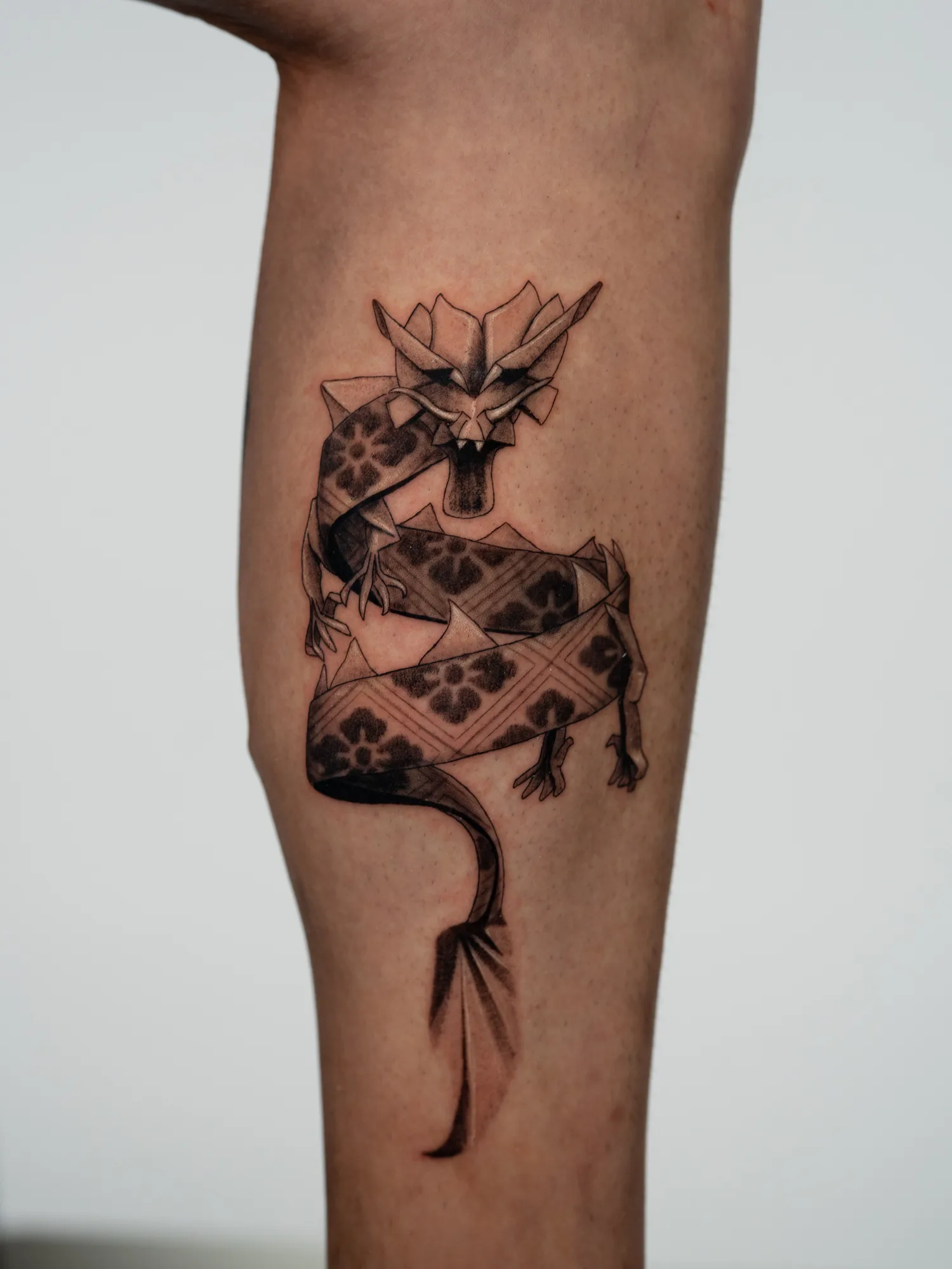 Black and grey tattoo of a dragon made of folded patterned paper, coiling down the side of a calf with its tail unrolling into a flat ribbon.