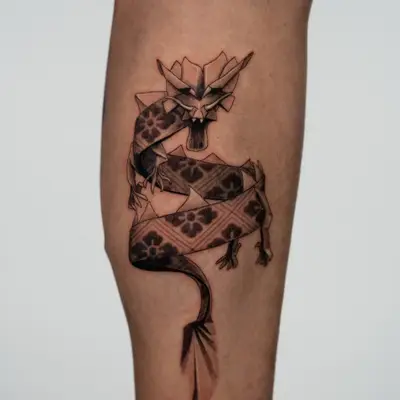 Black and grey tattoo of a dragon made of folded patterned paper, coiling down the side of a calf with its tail unrolling into a flat ribbon.
