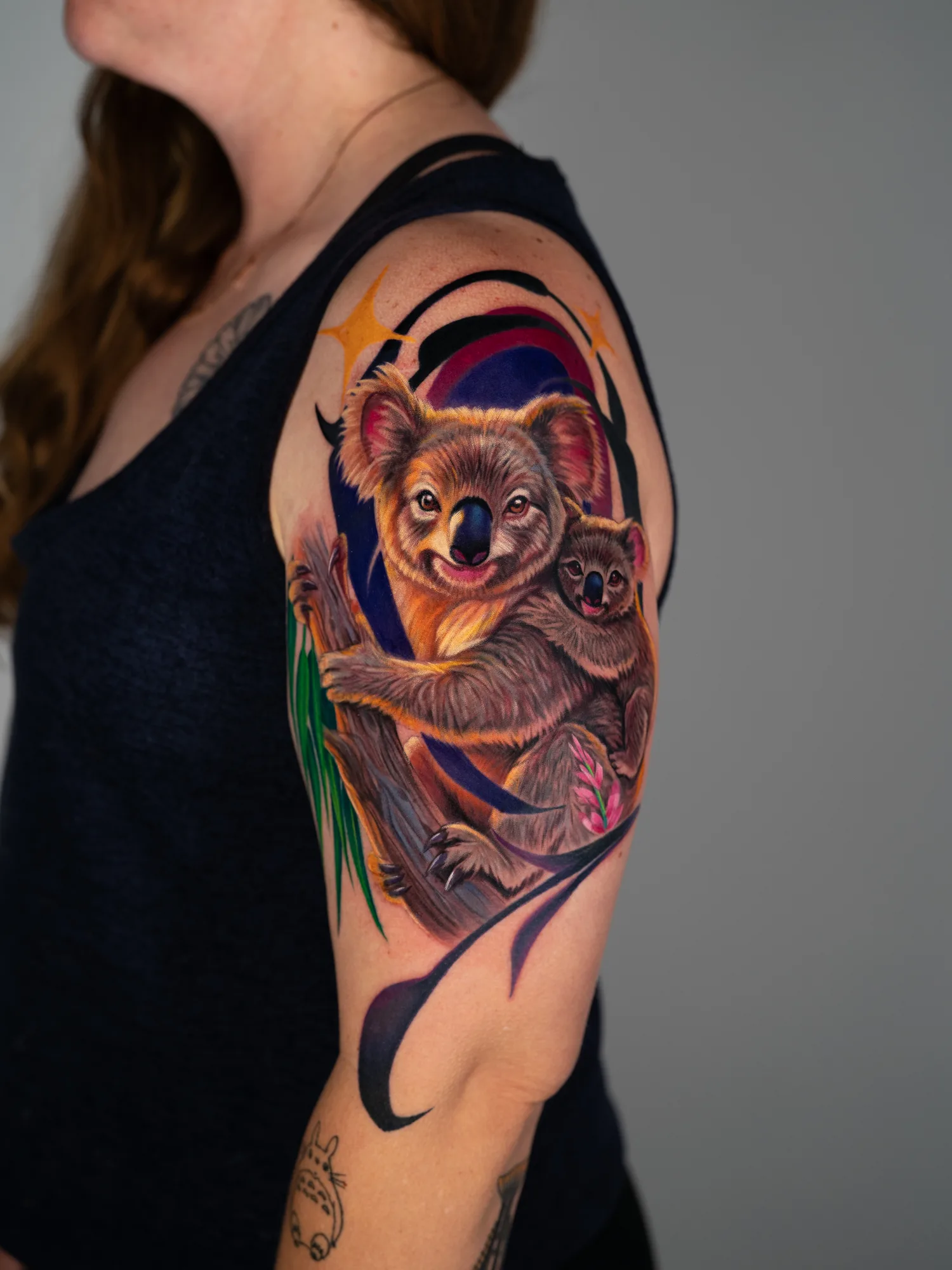 Full color realism tattoo of a mother koala with a joey on her back, holding a eucalyptus branch, against a purple and black graphic background on an upper arm.