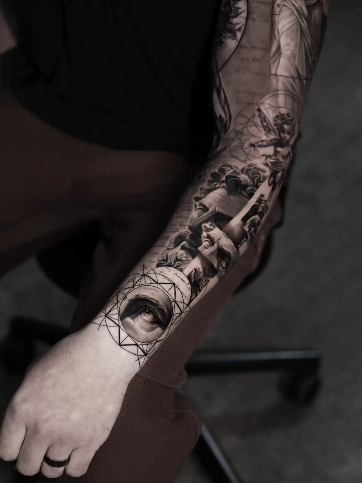 Black and grey full sleeve with a cherub, classical statue faces, handwritten script and a realistic eye set in a sacred geometry mandala at the wrist.