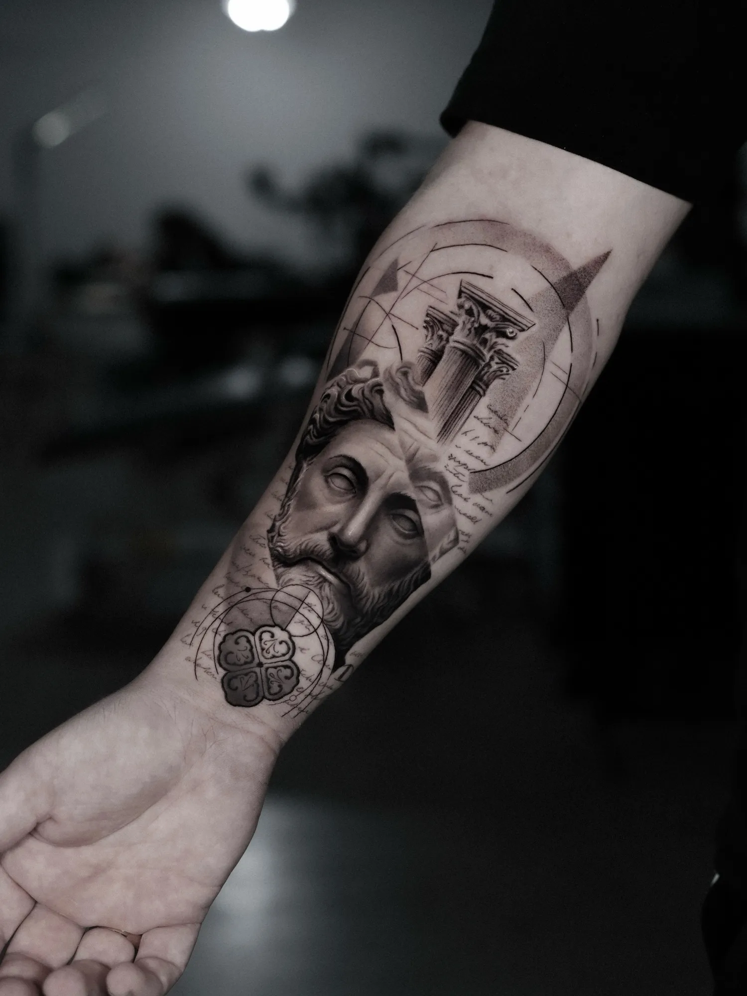 Black and grey forearm tattoo of a bearded marble bust below a Corinthian column capital, with geometric arcs and handwritten script.