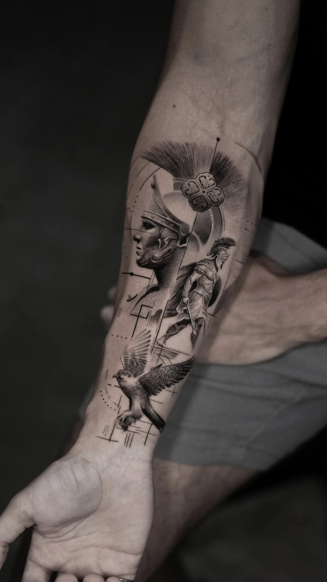 Black and grey forearm tattoo of a crested helmet in profile with a standing warrior figure and a bird in flight near the wrist.