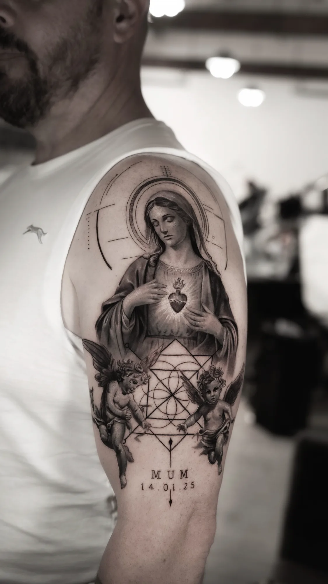 Black and grey half sleeve of the Virgin Mary with the Sacred Heart, a fine-line halo and arch, two cherubs and sacred geometry, with memorial lettering below.