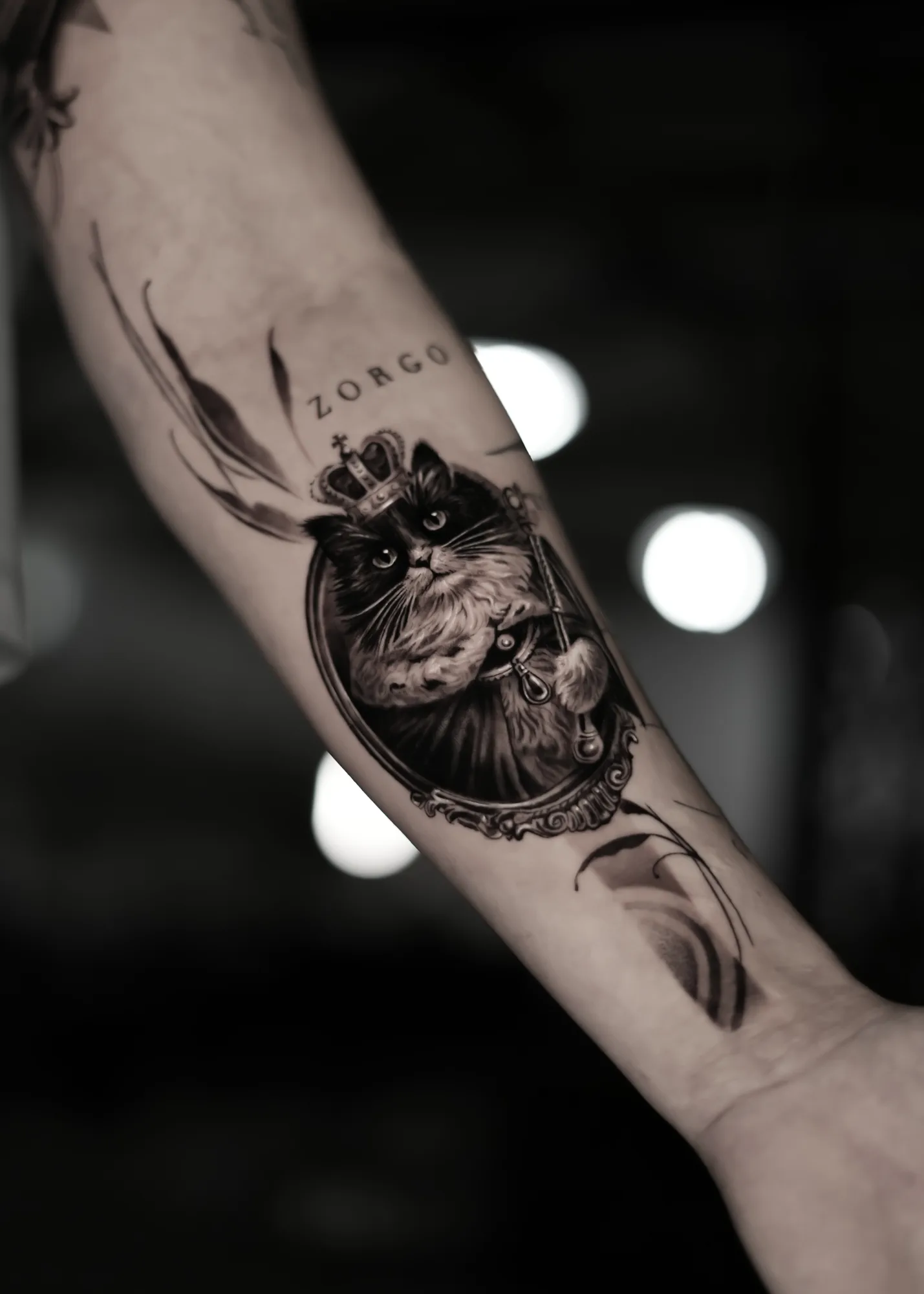 Black and grey forearm tattoo of a cat wearing a crown and holding a sceptre inside an ornate oval frame, with the name Zorgo lettered above.