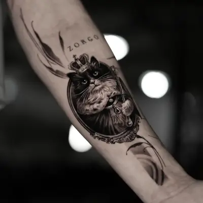 Black and grey forearm tattoo of a cat wearing a crown and holding a sceptre inside an ornate oval frame, with the name Zorgo lettered above.