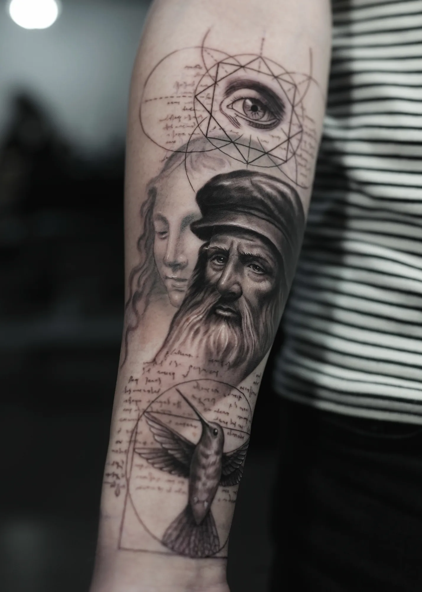 Black and grey forearm tattoo of Leonardo da Vinci with a woman's face behind him, an eye set in sacred geometry above and a hummingbird below.