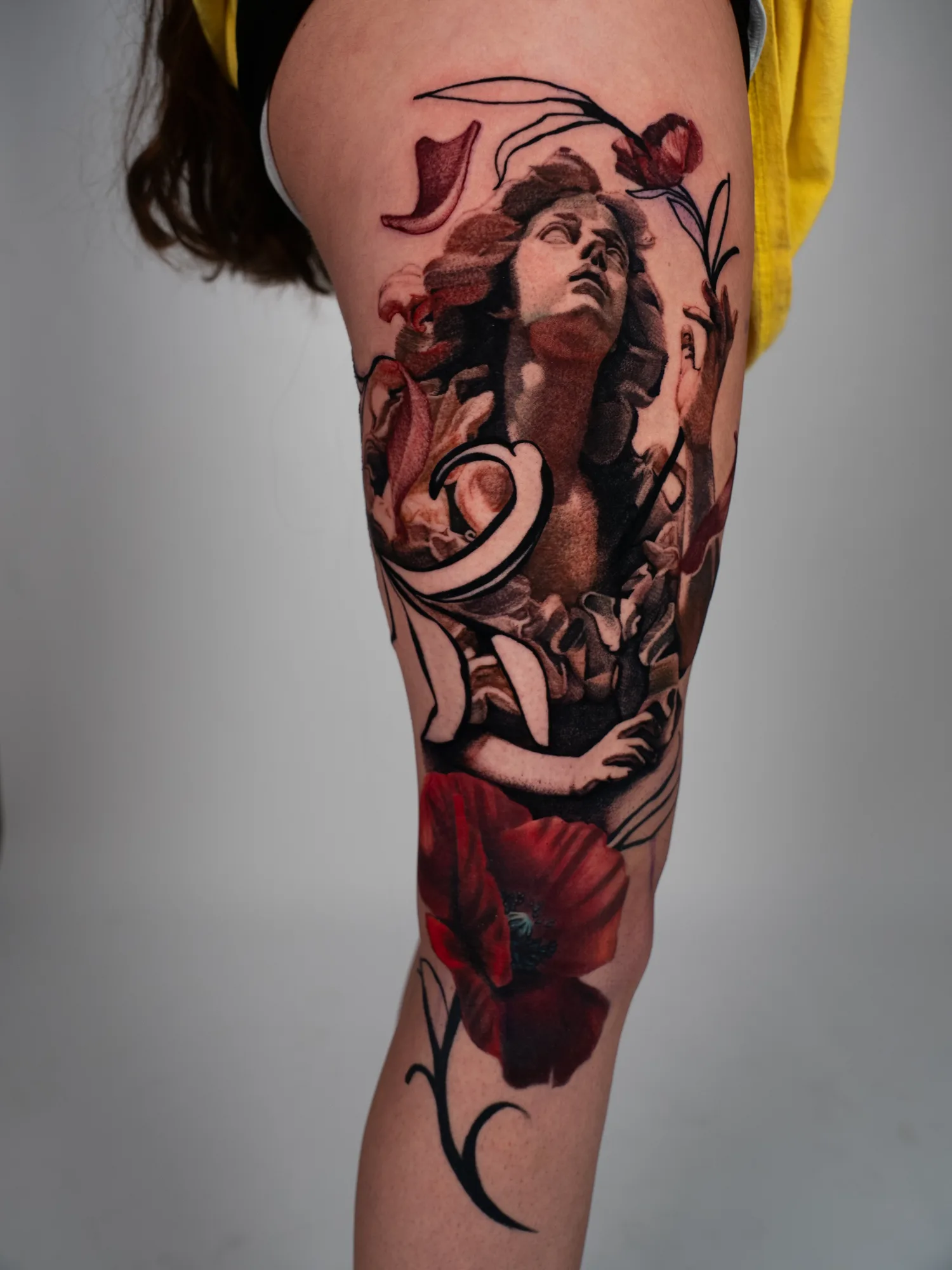 Classical statue figure looking upward, tattooed across a thigh in sepia and grey with dark red tulips, a large red poppy below the knee and graphic black line work running through the composition.