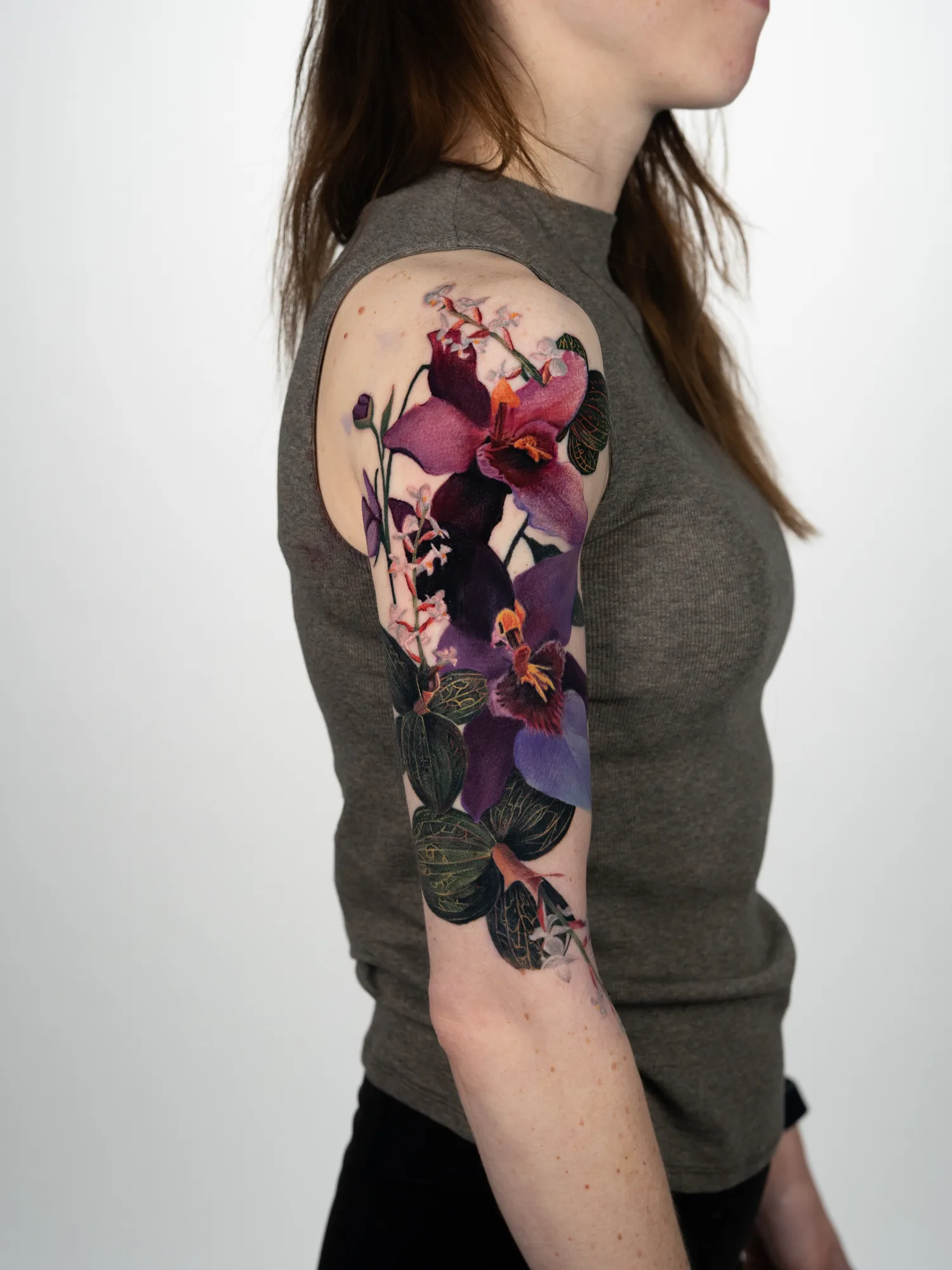 Color realism tattoo of purple and magenta jewel orchids with dark veined leaves, running down an upper arm.