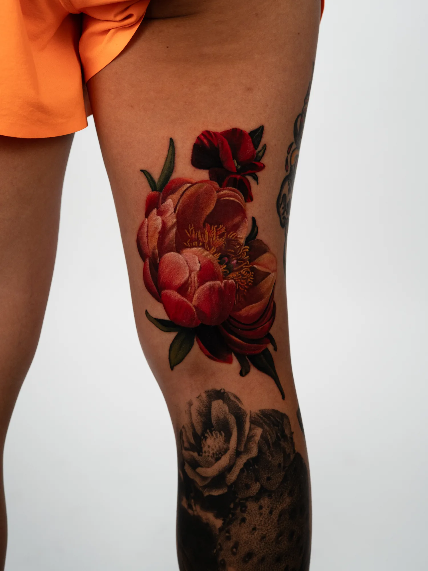 Color realism tattoo of coral and red peonies with a dark red iris above them, on a thigh just above the knee.