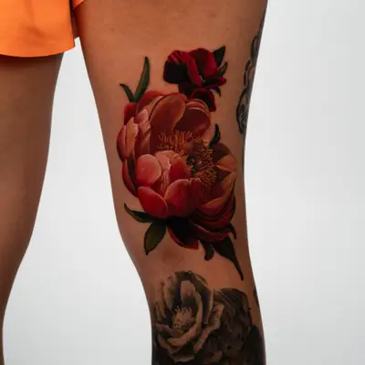 Color realism tattoo of coral and red peonies with a dark red iris above them, on a thigh just above the knee.