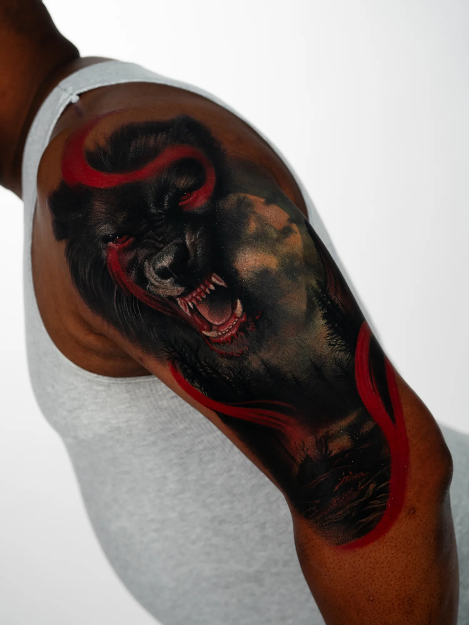 Snarling black werewolf with a silhouetted treeline behind it and red ribbon accents looping through, tattooed on an upper outer arm.