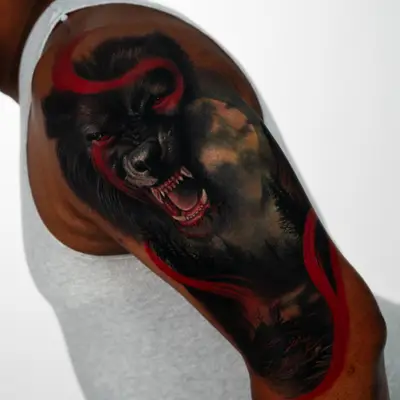Snarling black werewolf with a silhouetted treeline behind it and red ribbon accents looping through, tattooed on an upper outer arm.
