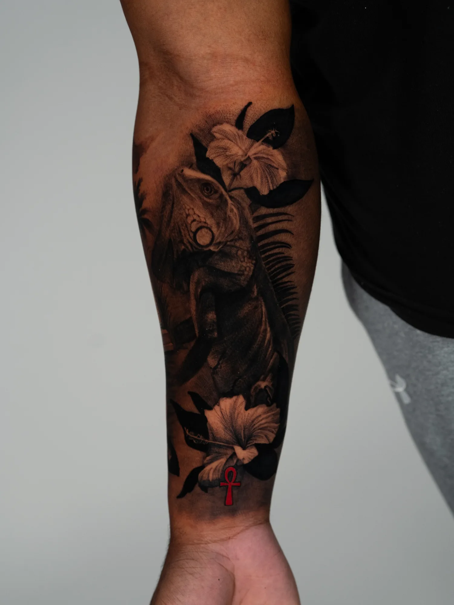 Iguana climbing among hibiscus flowers and palm fronds, wrapping a forearm in black and grey, with a small red ankh near the wrist.