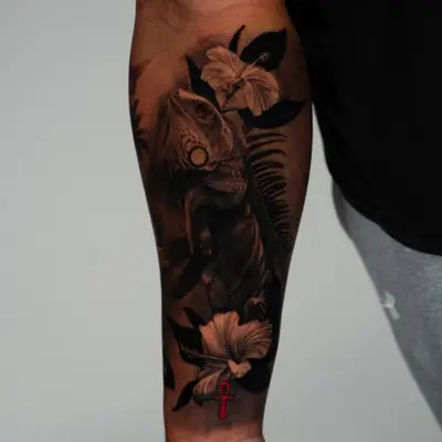 Iguana climbing among hibiscus flowers and palm fronds, wrapping a forearm in black and grey, with a small red ankh near the wrist.