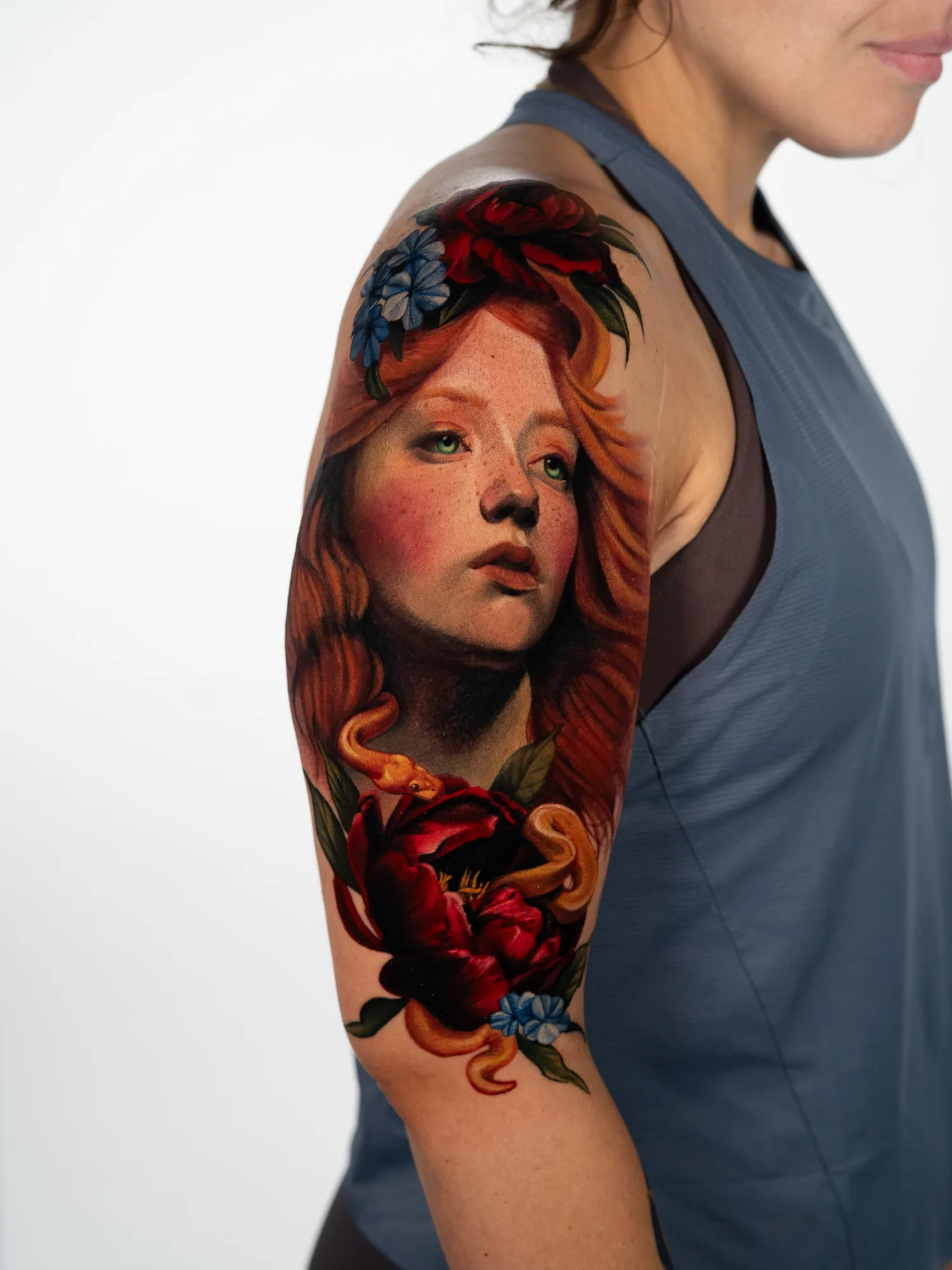 Color realism portrait of a red-haired woman with green eyes on an upper arm, crowned with red peonies and blue cornflowers, a gold snake winding down through the flowers.