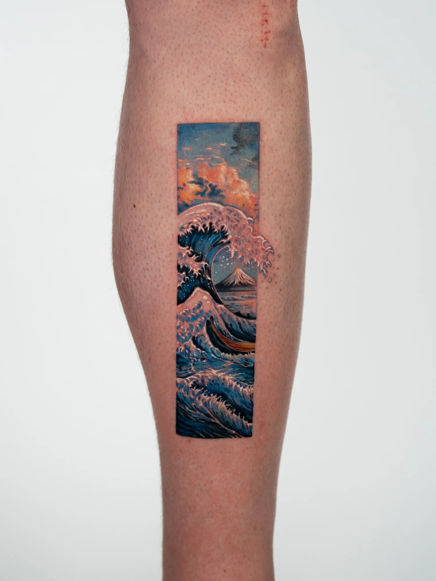 Hokusai's Great Wave with Mount Fuji behind it, tattooed in a vertical frame on the side of a calf, with foam breaking out past the border.