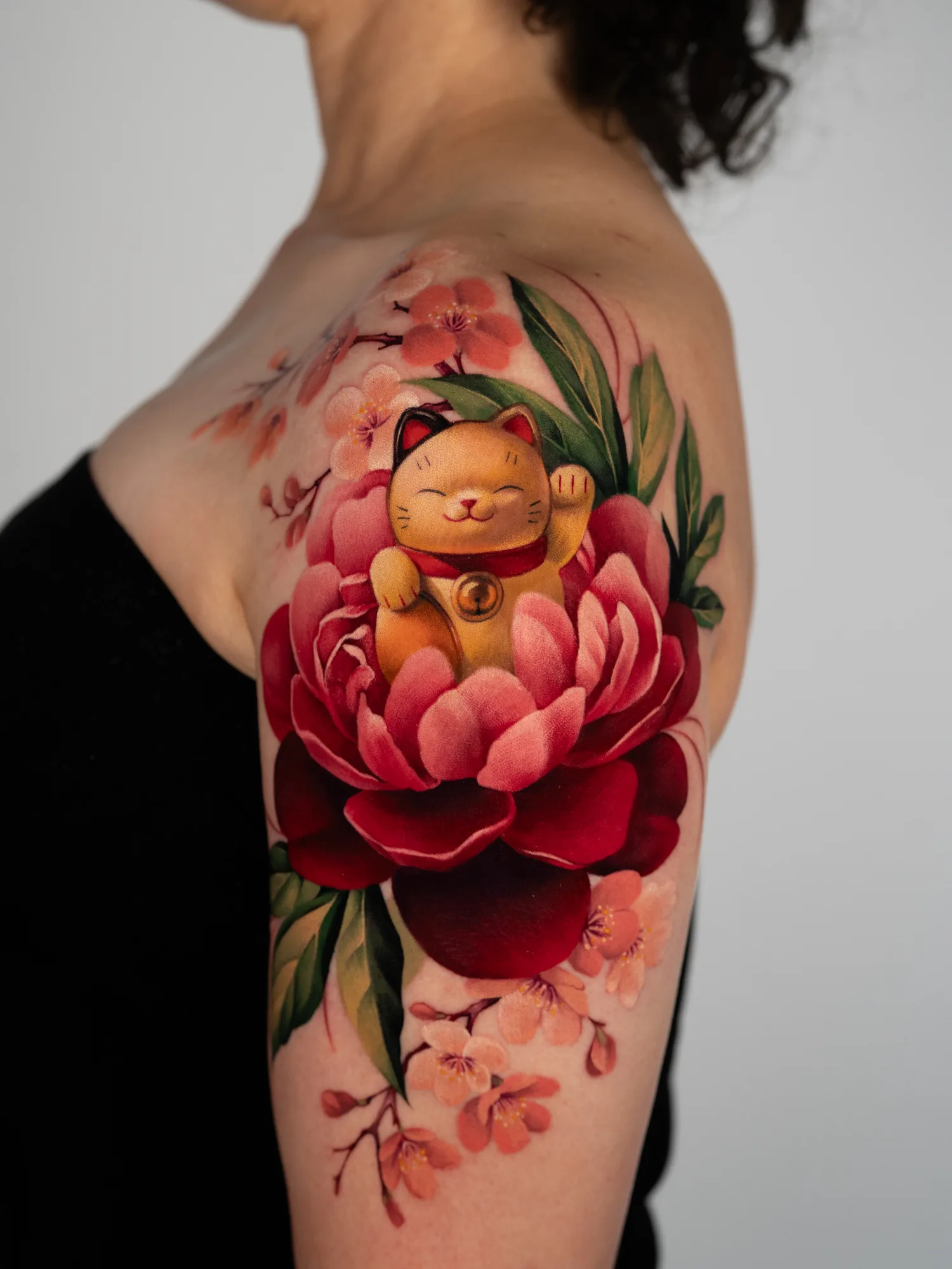 Color tattoo on the shoulder and upper arm: a golden Maneki-Neko lucky cat sitting inside a red and pink peony, with sakura branches running toward the collarbone and down the arm.