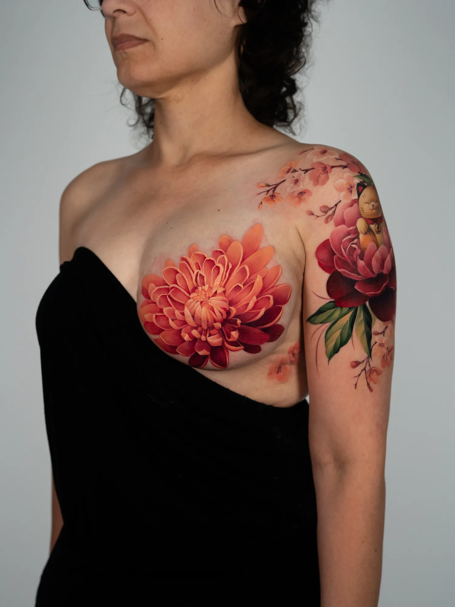 Color tattoo on the chest: a large orange and crimson chrysanthemum with a few sakura blossoms below it, beside a lucky cat and peony piece on the shoulder.