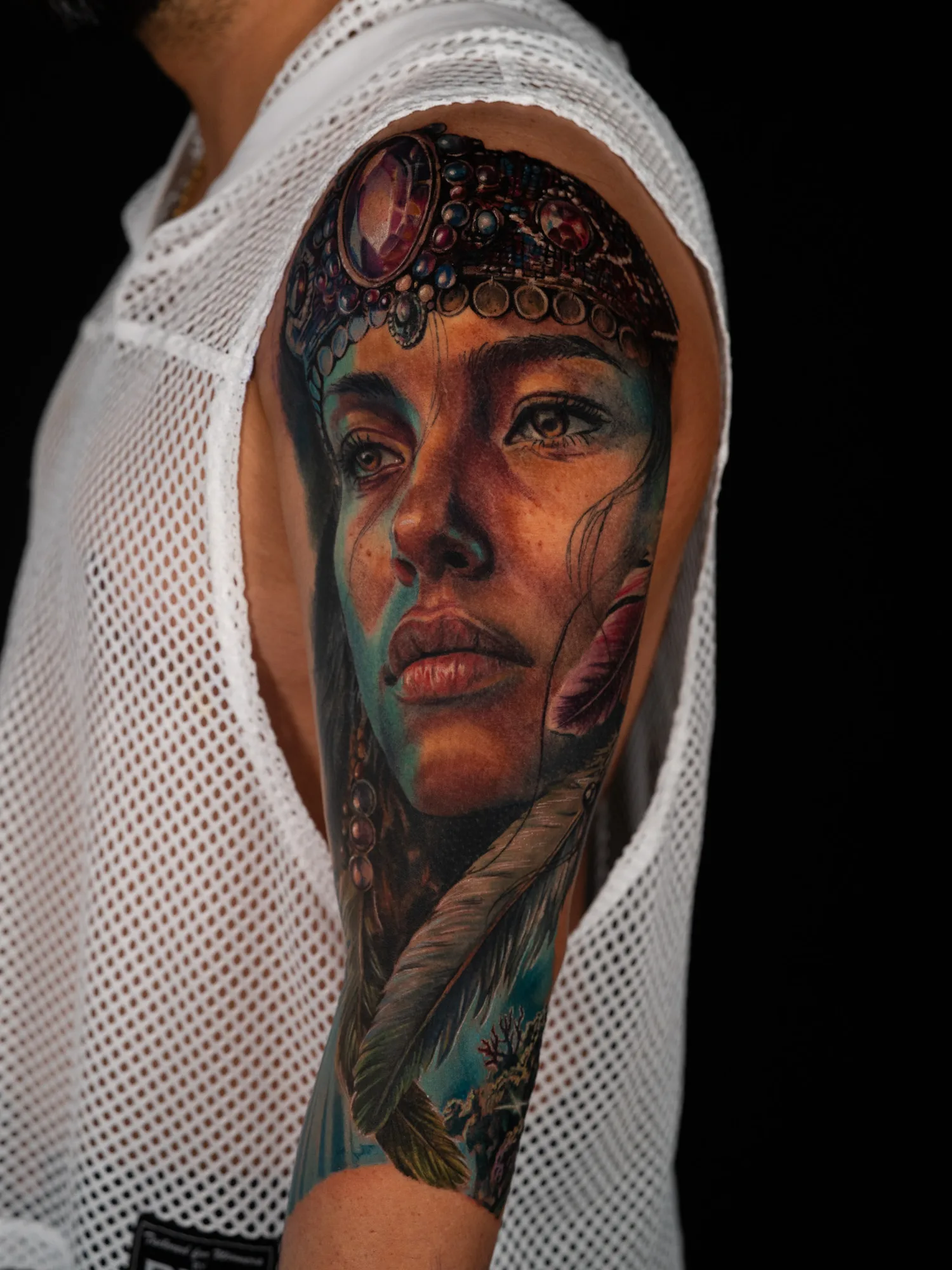 Work-in-progress color realism tattoo on the outer upper arm: a woman's face in a jeweled headdress with feathers and beaded strands, teal shadows on her face, and coral starting below.