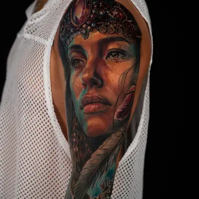 Work-in-progress color realism tattoo on the outer upper arm: a woman's face in a jeweled headdress with feathers and beaded strands, teal shadows on her face, and coral starting below.