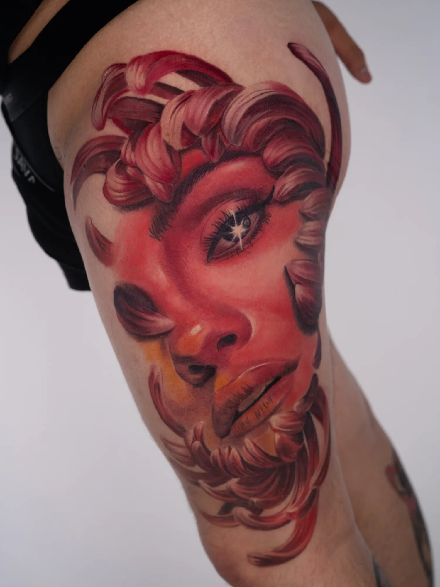 Color realism tattoo on the upper thigh: Selena Gomez portrait in deep reds and pinks framed by curling chrysanthemum petals in place of hair, with a starburst highlight in her eye.