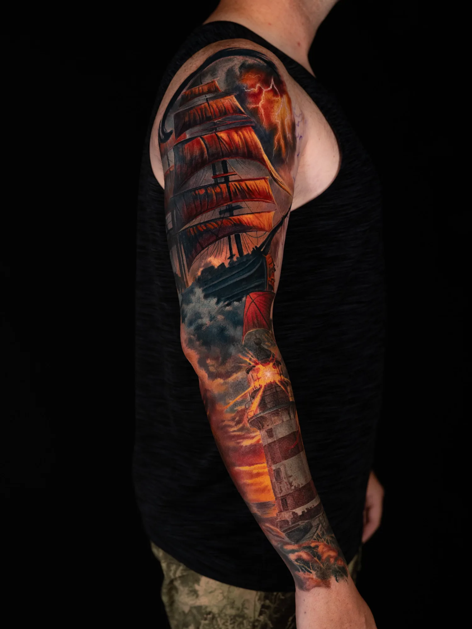 Color realism full sleeve: a tall ship with orange sails in a lightning storm on the upper arm, with clouds opening into a sunset over a red and white lighthouse on the forearm.