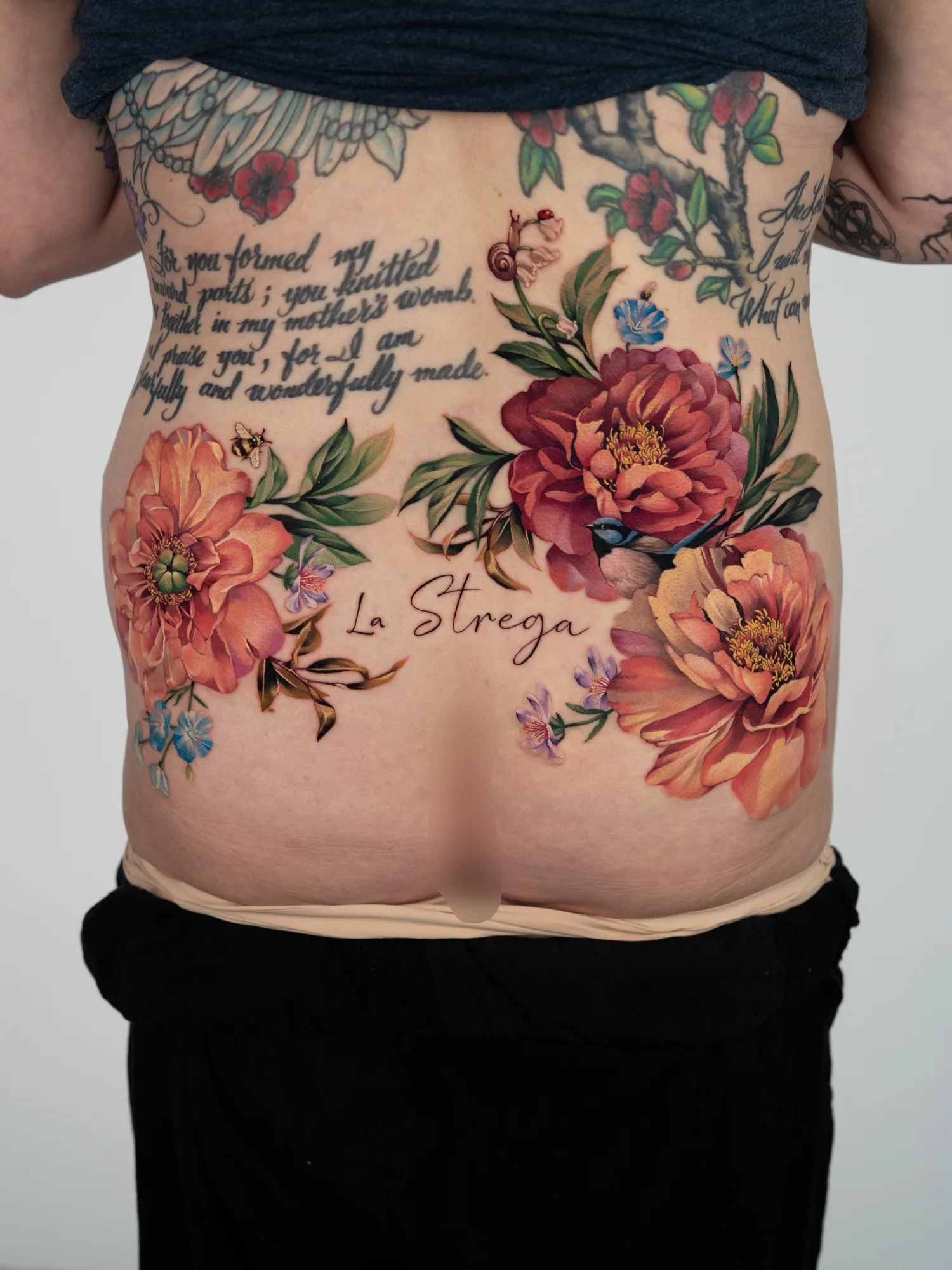 Color floral tattoo across the lower back: two large coral peonies on each side with a bee, a snail, a small blue bird and blue and lilac blossoms, and the words La Strega in flowing script at the center.