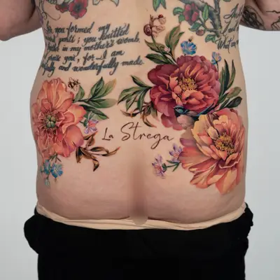 Color floral tattoo across the lower back: two large coral peonies on each side with a bee, a snail, a small blue bird and blue and lilac blossoms, and the words La Strega in flowing script at the center.