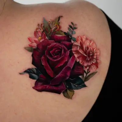 Color realism tattoo on the upper back over the shoulder blade: a deep red rose with a pink carnation, orange honeysuckle, a small pink flower and dark green leaves.