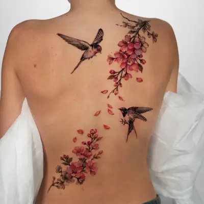 Black and grey tattoo of two swallows in flight with pink cherry blossom branches and falling petals, running diagonally across the back from the right shoulder to the lower left back.
