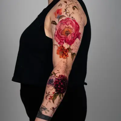 Color realism floral sleeve in progress on the outer arm: a large pink peony and orange blooms on the upper arm, a deep red dahlia and lavender anemone at the elbow, with unshaded line work for flowers still to come.