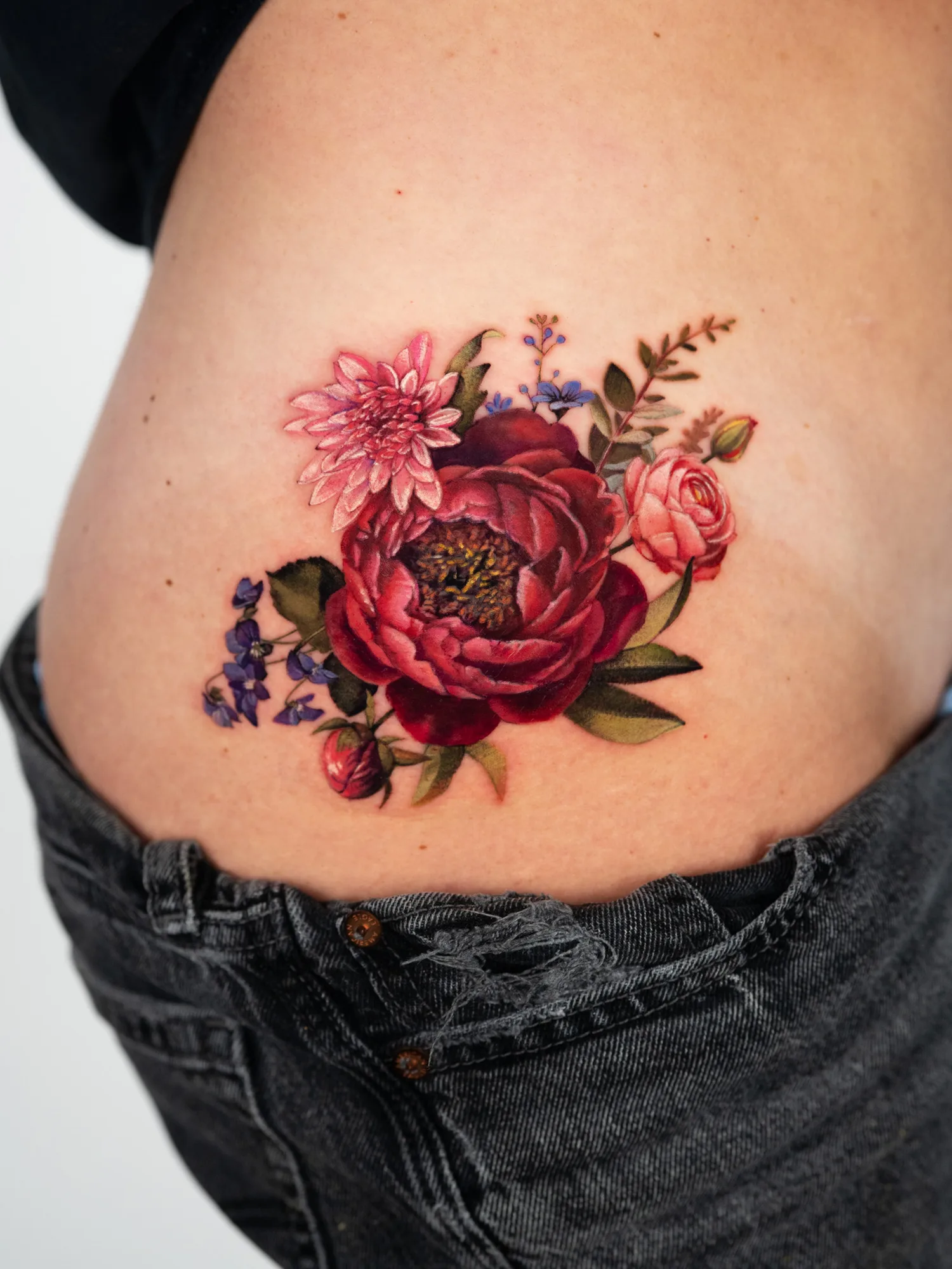 Color realism tattoo on the hip of a deep red peony with a pink dahlia, a pink ranunculus, purple violets, a peony bud and green leaves.