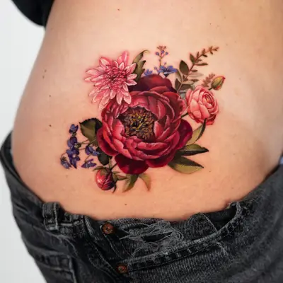Color realism tattoo on the hip of a deep red peony with a pink dahlia, a pink ranunculus, purple violets, a peony bud and green leaves.