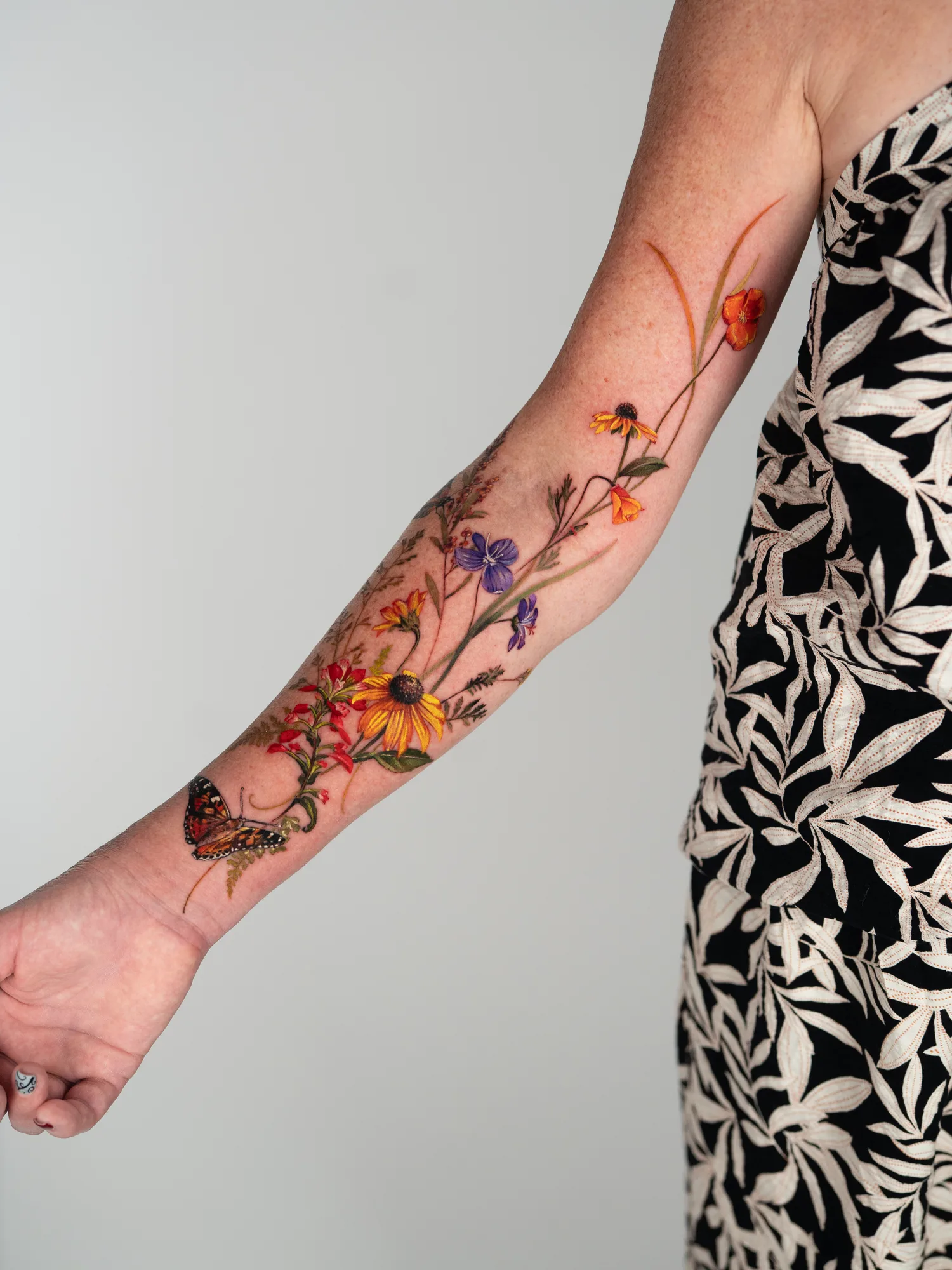 Color tattoo of loose wildflowers on the inner arm from the wrist to above the elbow: red Indian paintbrush, black-eyed Susans, purple violets, orange poppies, grasses, ferns and a painted lady butterfly near the wrist.