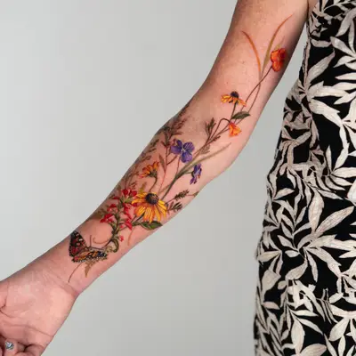 Color tattoo of loose wildflowers on the inner arm from the wrist to above the elbow: red Indian paintbrush, black-eyed Susans, purple violets, orange poppies, grasses, ferns and a painted lady butterfly near the wrist.