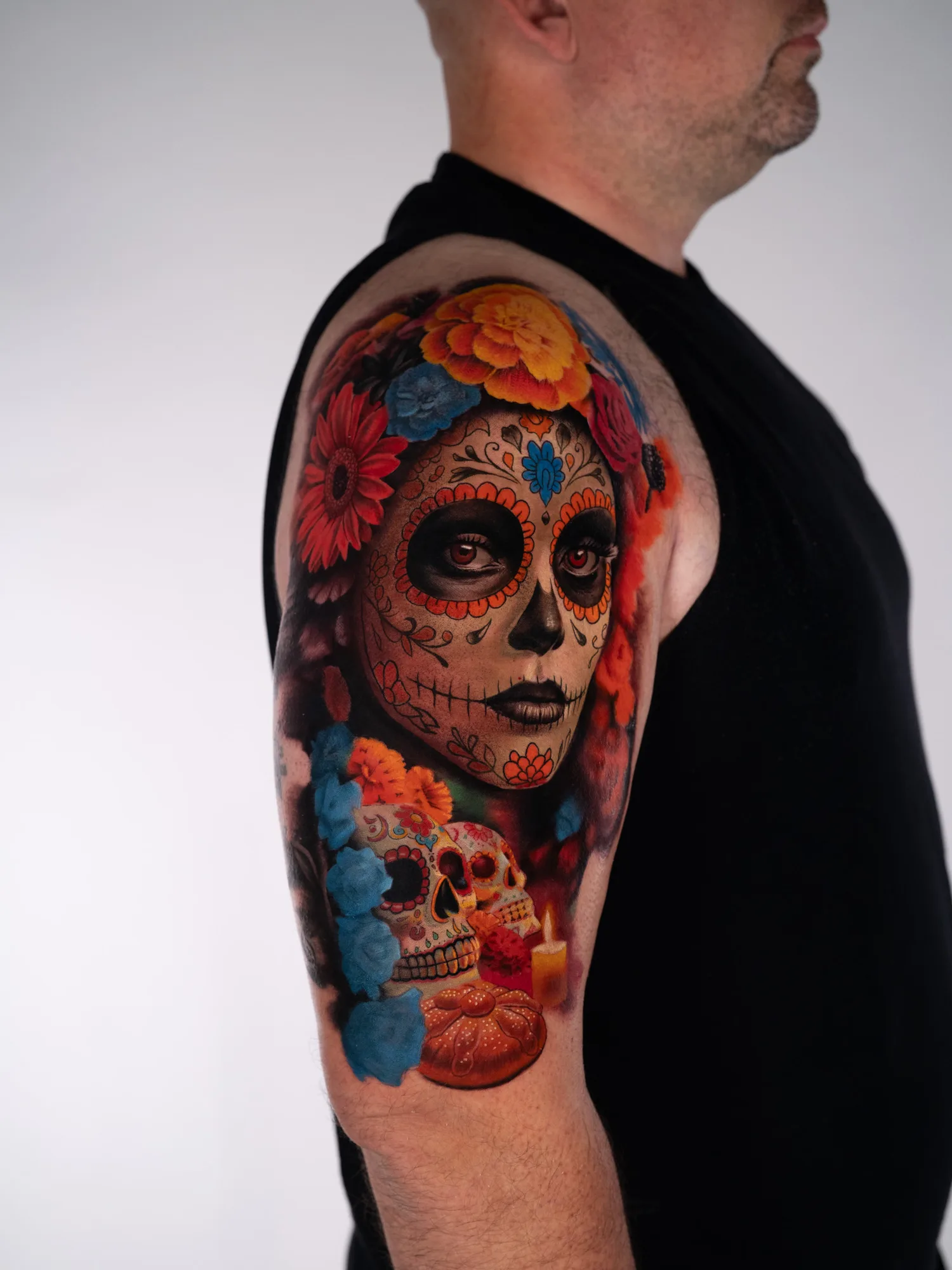 Color realism tattoo in progress on the outer upper arm: a woman in Day of the Dead sugar skull makeup crowned with marigolds and roses, with two sugar skulls, pan de muerto and a lit candle below.
