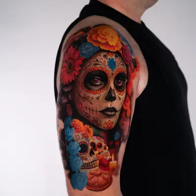 Color realism tattoo in progress on the outer upper arm: a woman in Day of the Dead sugar skull makeup crowned with marigolds and roses, with two sugar skulls, pan de muerto and a lit candle below.