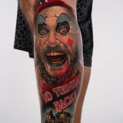 Color realism tattoo on the lower leg of Captain Spaulding, the clown from House of 1000 Corpses, laughing under a stars-and-stripes top hat, with a No Turning Back! sign and a lit house with a graveyard below.