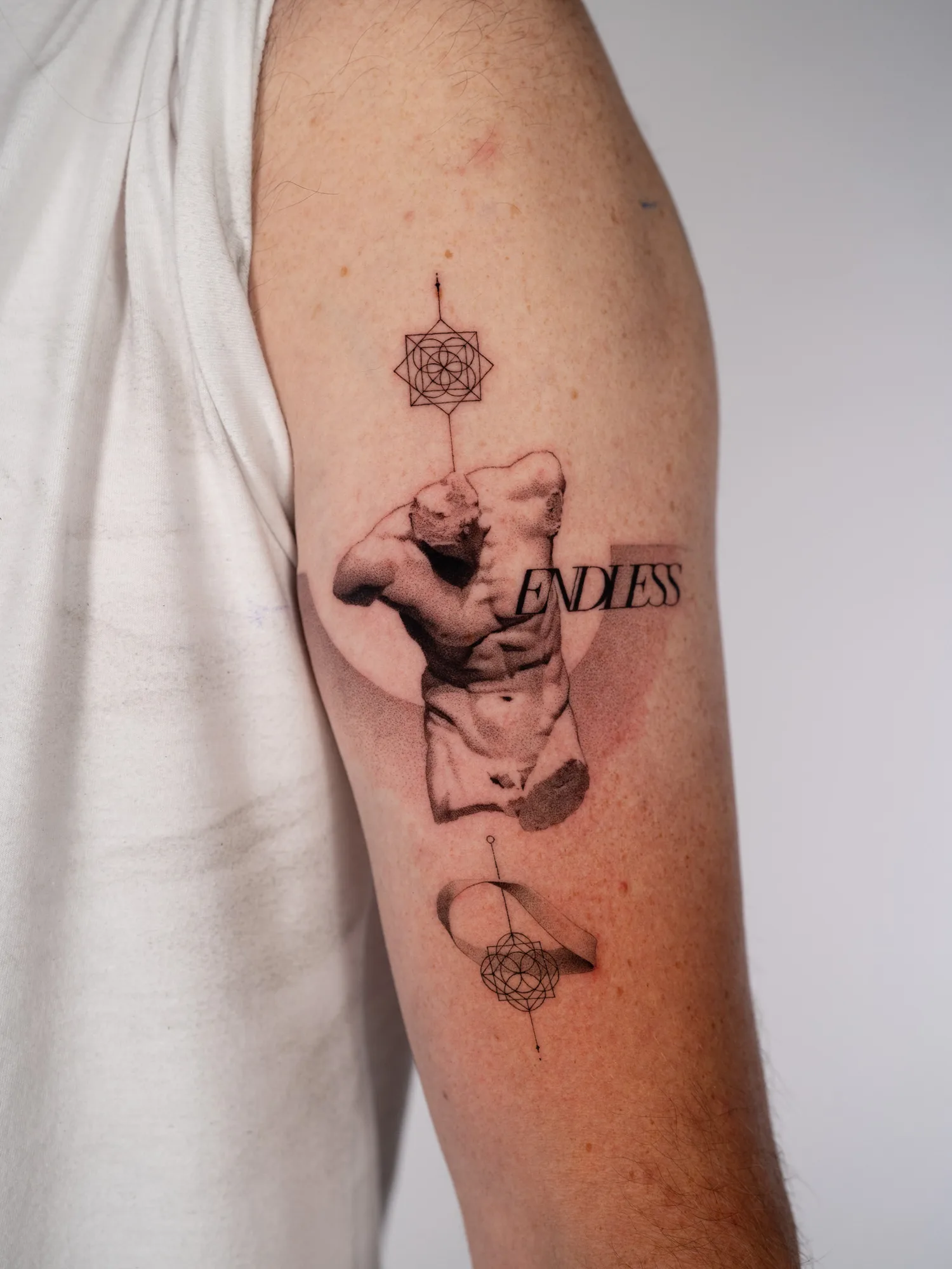 Black and grey fine-line tattoo on the back of a man's upper arm: a classical marble statue torso with the word ENDLESS set across it, a soft grey half-circle behind, and two geometric sacred-geometry marks on a vertical line above and below, the lower one wrapped by a Möbius strip.