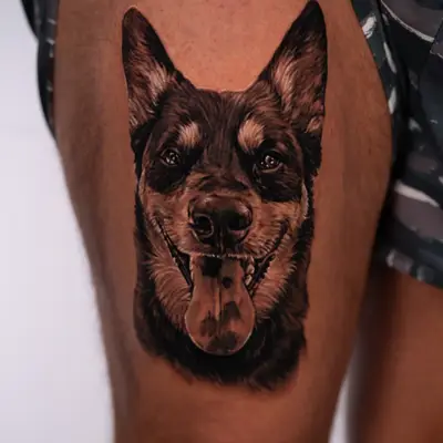 Black and grey realistic portrait of a dog with tall pointed ears and an open-mouthed, tongue-out smile, tattooed on the front of the right thigh.
