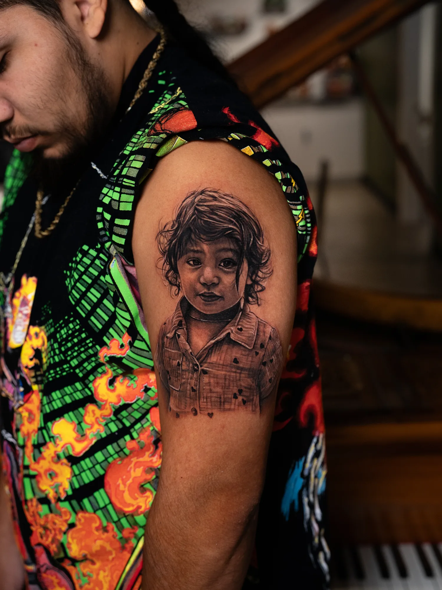 Black and grey realistic portrait of a young child with loose curly hair, wearing a collared shirt printed with small hearts, tattooed on the outer upper arm of a man.