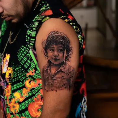 Black and grey realistic portrait of a young child with loose curly hair, wearing a collared shirt printed with small hearts, tattooed on the outer upper arm of a man.